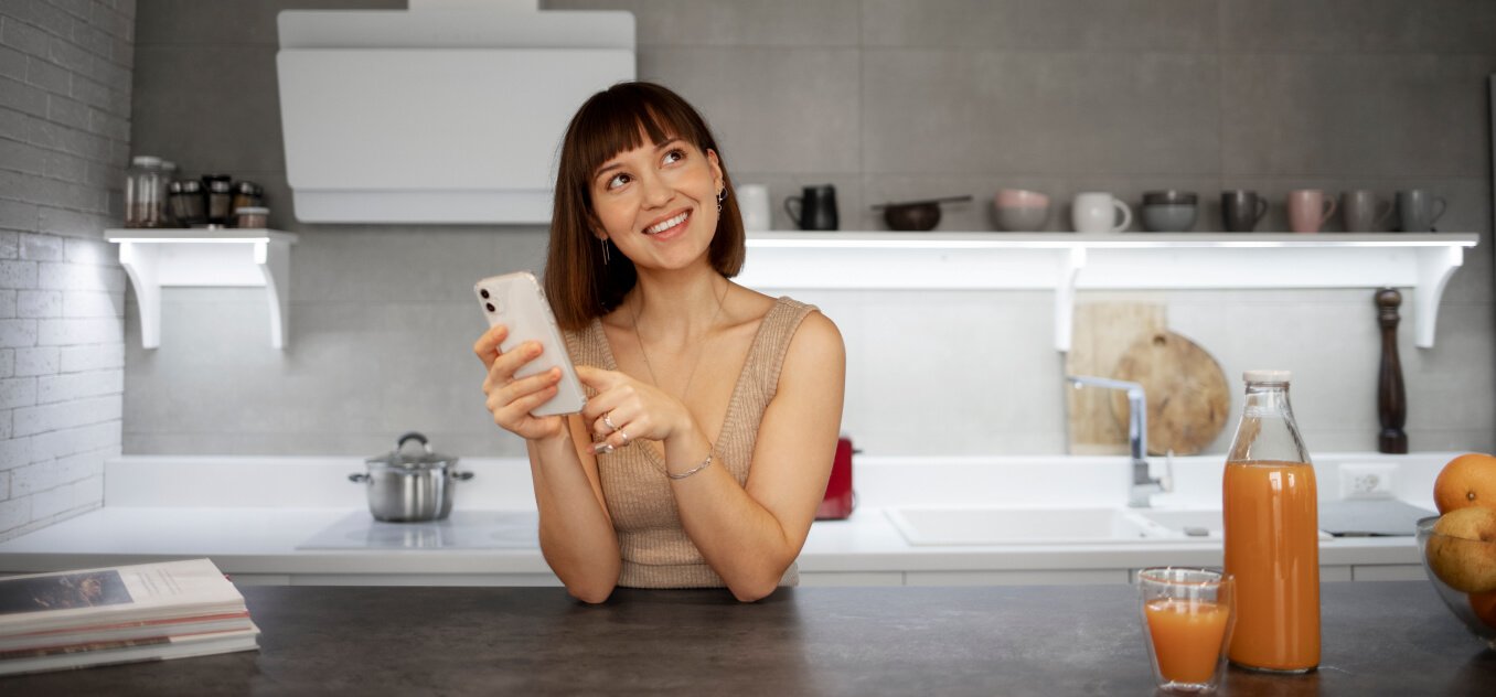a smiling woman sitting in the kitchen uses a smartphone