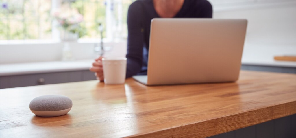 google nest mini umieszczony na biurku, w tle osoba korzystająca z laptopa