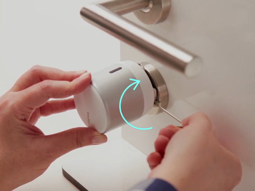 An image showing hands installing the Tedee GO smart lock onto a European cylinder. The person's left hand is holding the smart lock in place while the right hand is using a screwdriver to fix a screw, with a curved arrow symbol indicating the tightening direction.
