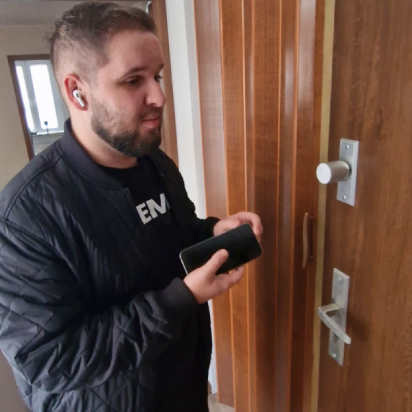 A man is standing by a door, interacting with a Tedee PRO smart lock mounted on the door's surface. He holds a smartphone in one hand, using it to control the smart lock. The man is Adrian Now, an online influencer with a visual impairment who specializes in assistive technologies.